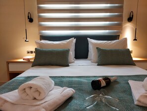 Habitación de hotel con cama, almohadas, mesita de noche, lámpara, botella y toalla.