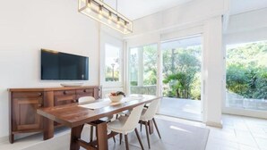 A modern dining area with a wooden table, white chairs, a television, and a large window offering a view of greenery. - Hostdomus - Queen Suite (Marina di Andora)
