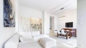 A modern living room with a sectional sofa, a dining area with chairs, a TV, and a staircase leading to another level. - Hostdomus - Queen Suite (Marina di Andora)