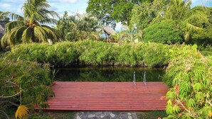Um deck de madeira com um caminho de pedras que leva a uma estrutura com telhado de palha, cercada por vegetação exuberante e um corpo d'água calmo.