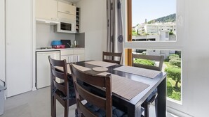 A compact kitchen with modern appliances, a dining table with chairs, and a large window offering a view of buildings and greenery. - Flat with a swimming pool and a balcony with mountain views (Grasse)