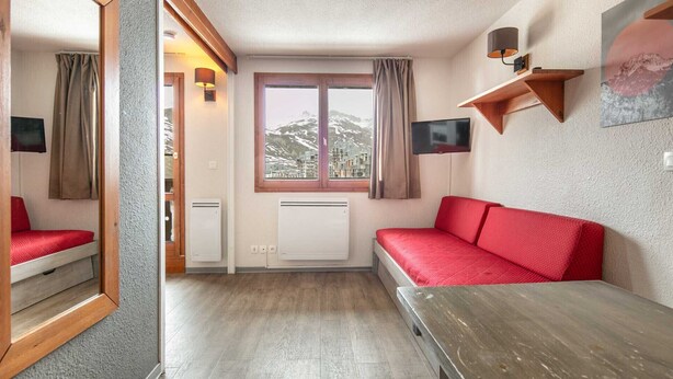 Ein Zimmer mit einem roten Sofa, einem Holztisch, einem Fenster mit Blick auf Gebäude, einem an der Wand montierten Fernseher und einer Lampe.