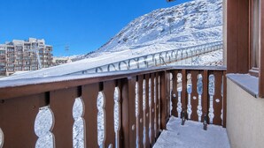 Ein mit Schnee bedeckter Balkon mit Metallgeländer, ein strahlend blauer Himmel und eine gebirgige Landschaft im Hintergrund.