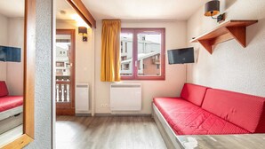 Een kamer met een rode bank, een raam met gele gordijnen, een aan de muur bevestigde televisie en een radiator.