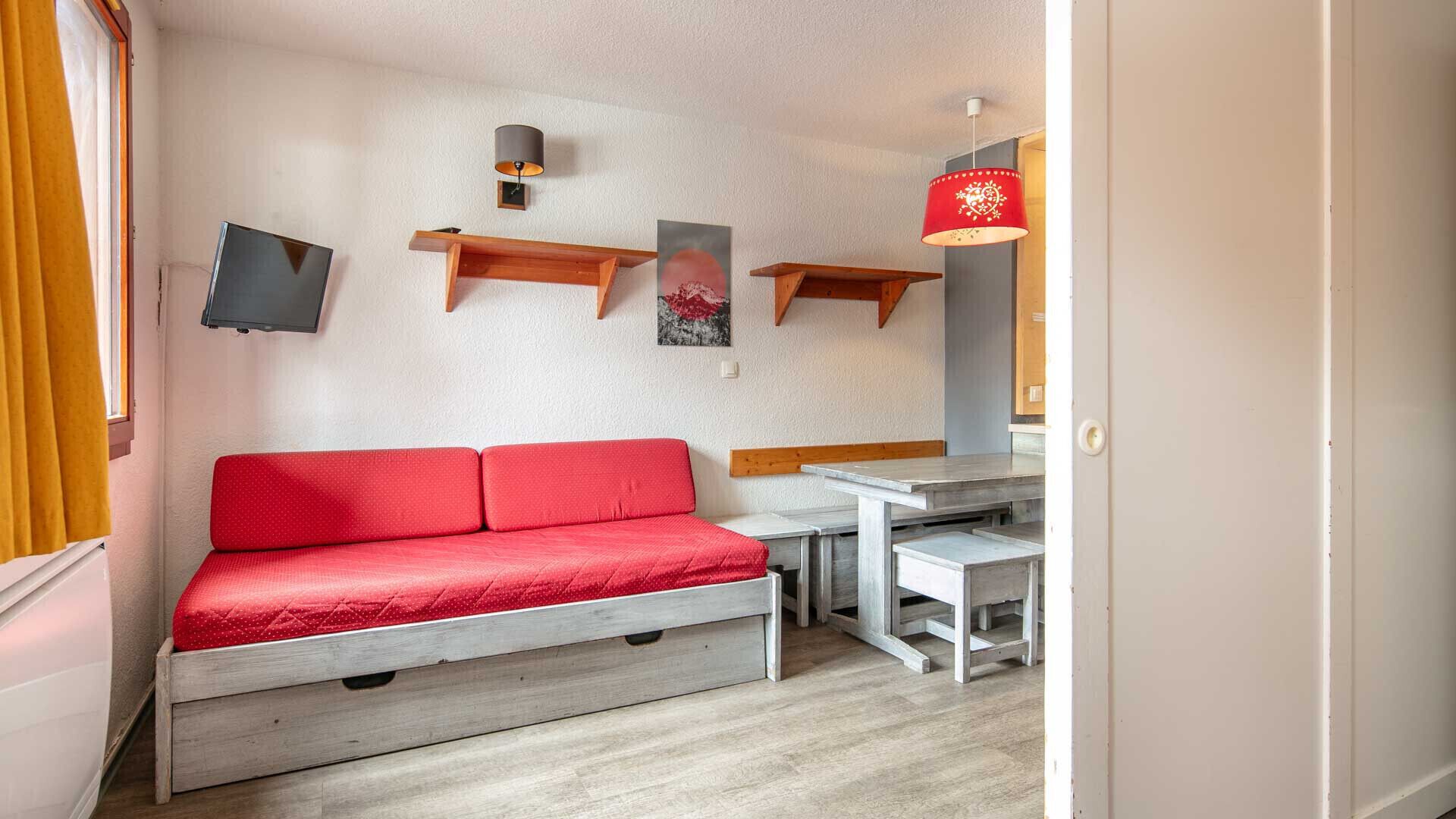 A room with a red sofa, a small table, a TV mounted on the wall, and a red hanging lamp.