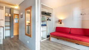 Een knus kamertje met een rode bank, een houten bankje, een kast en een kleine kitchenette.