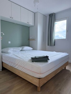 A bedroom with a bed, wooden headboard, white cabinets, a window with curtains, and a light fixture. - Surf & Sun - Cozy One-Bedroom Apartment Near the Ocean (Soustons)