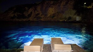 A nighttime swimming pool with clear water, two lounge chairs, and a rocky cliff in the background. - Milos Boathouse-Only one in Milos with sea view on both sides & stunning sunset  (Areti)