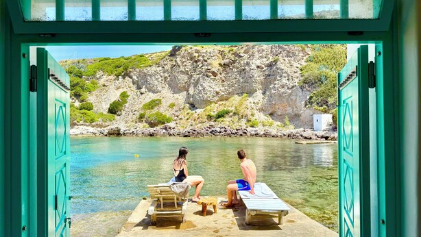 Uitzicht door open deuren op een zwembadgedeelte met twee lounge fauteuils en een tafeltje, met zicht op helder water en rotsachtig terrein.