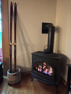 Ein Kaminofen mit sichtbaren Flammen, ein Paar Ski, die an einer Wand lehnen, und ein Korbwägelchen auf dem Boden.