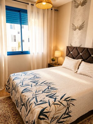 A bedroom with a bed, curtains, a nightstand, a lamp, and a wall with a decorative pattern. - Pearl Andalusia Seaside Oasis (El Mansouria)
