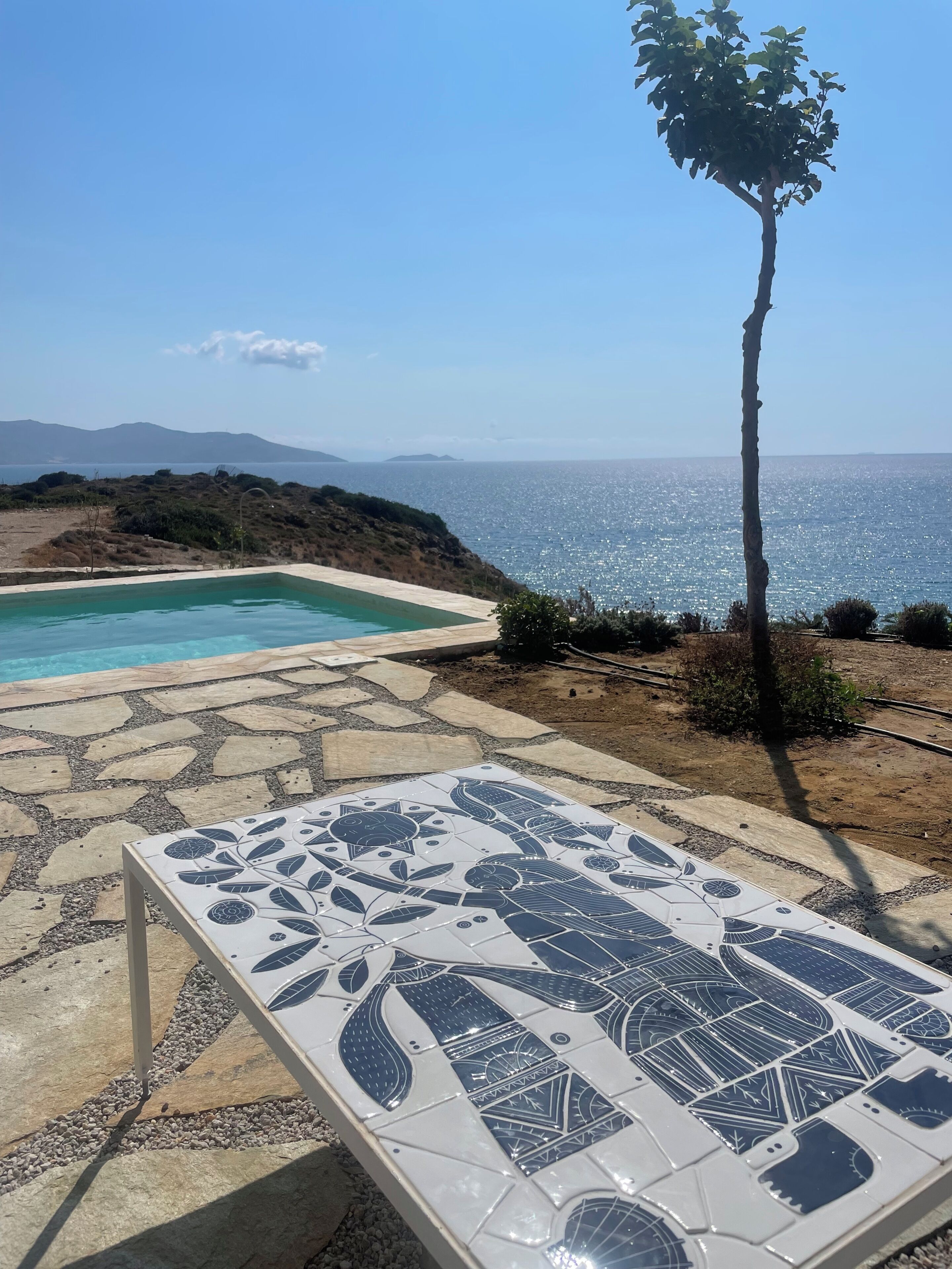 Una piscina con solárium de baldosas, una palmera y vistas al mar y las montañas.