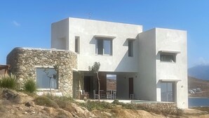 Une maison moderne à deux étages, dotée d’une entrée avec un mur en pierre, de grandes fenêtres et offrant une vue sur la mer.