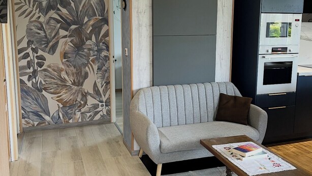 Ein modernes Wohnzimmer mit einem grauen Sofa, einem Holztisch, einer Wand mit Muster und einer eingebauten Mikrowelle.