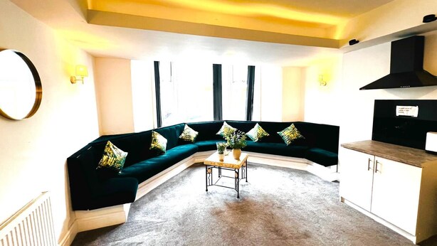Ein Wohnzimmer mit einem dunkelgrünen Sofa, einem kleinen Couchtisch und einer Küchenzeile mit einer schwarzen Dunstabzugshaube.