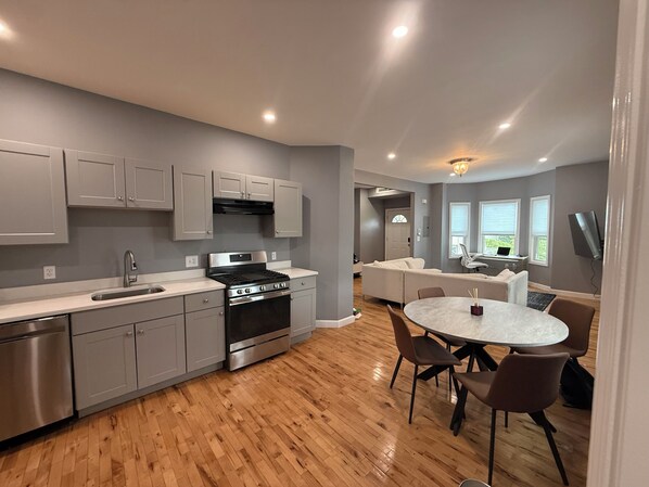 Una cocina moderna con electrodomésticos de acero inoxidable, un comedor con una mesa redonda y sillas, y una sala de estar con sofá y televisor.