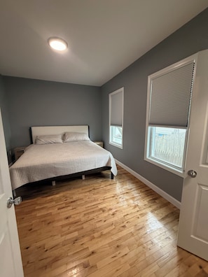 Un dormitorio con cama, piso de madera, ventana con persianas y puerta.