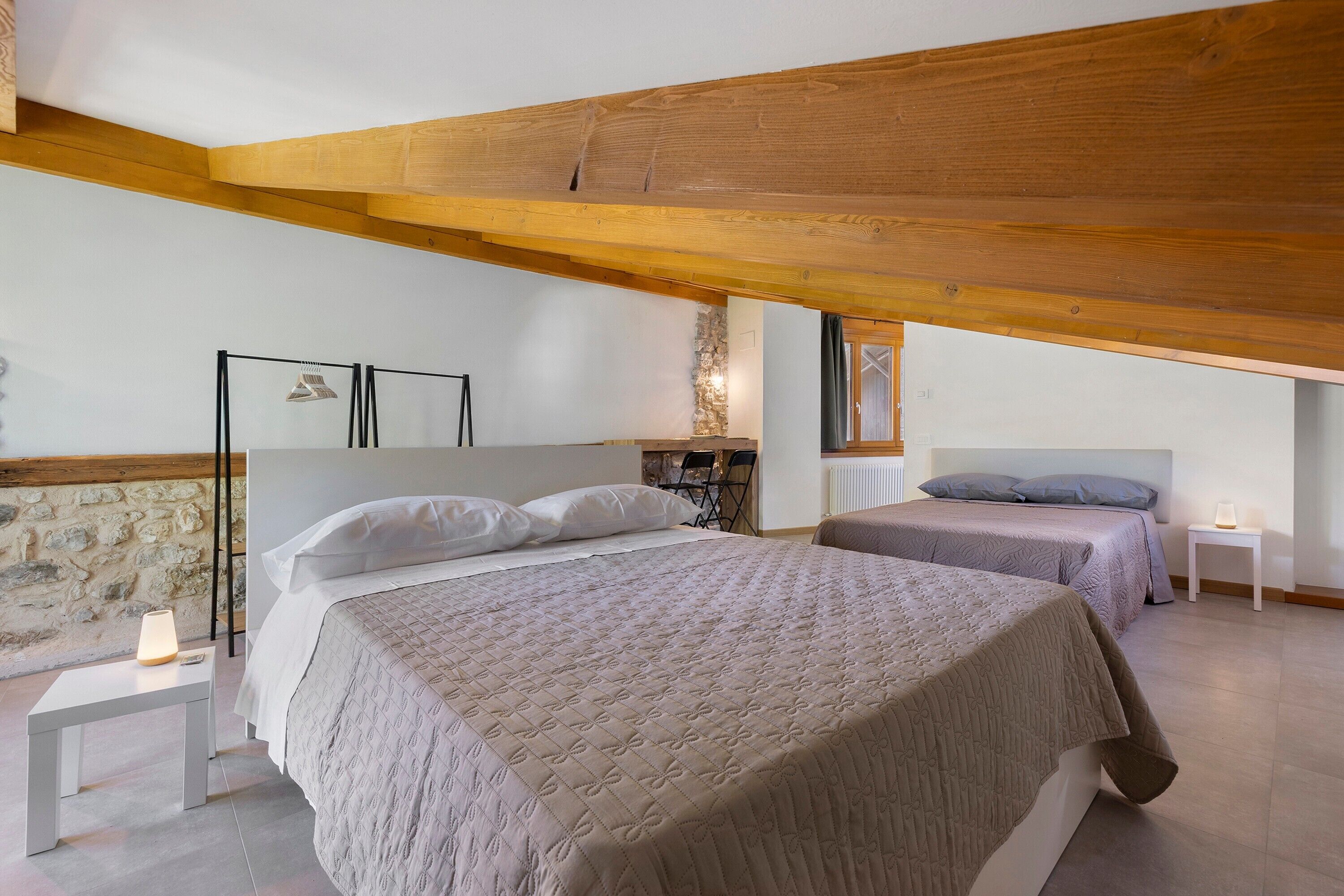 Un dormitorio con dos camas, una pared de piedra, un travesaño de techo de madera y una mesita con una lámpara.