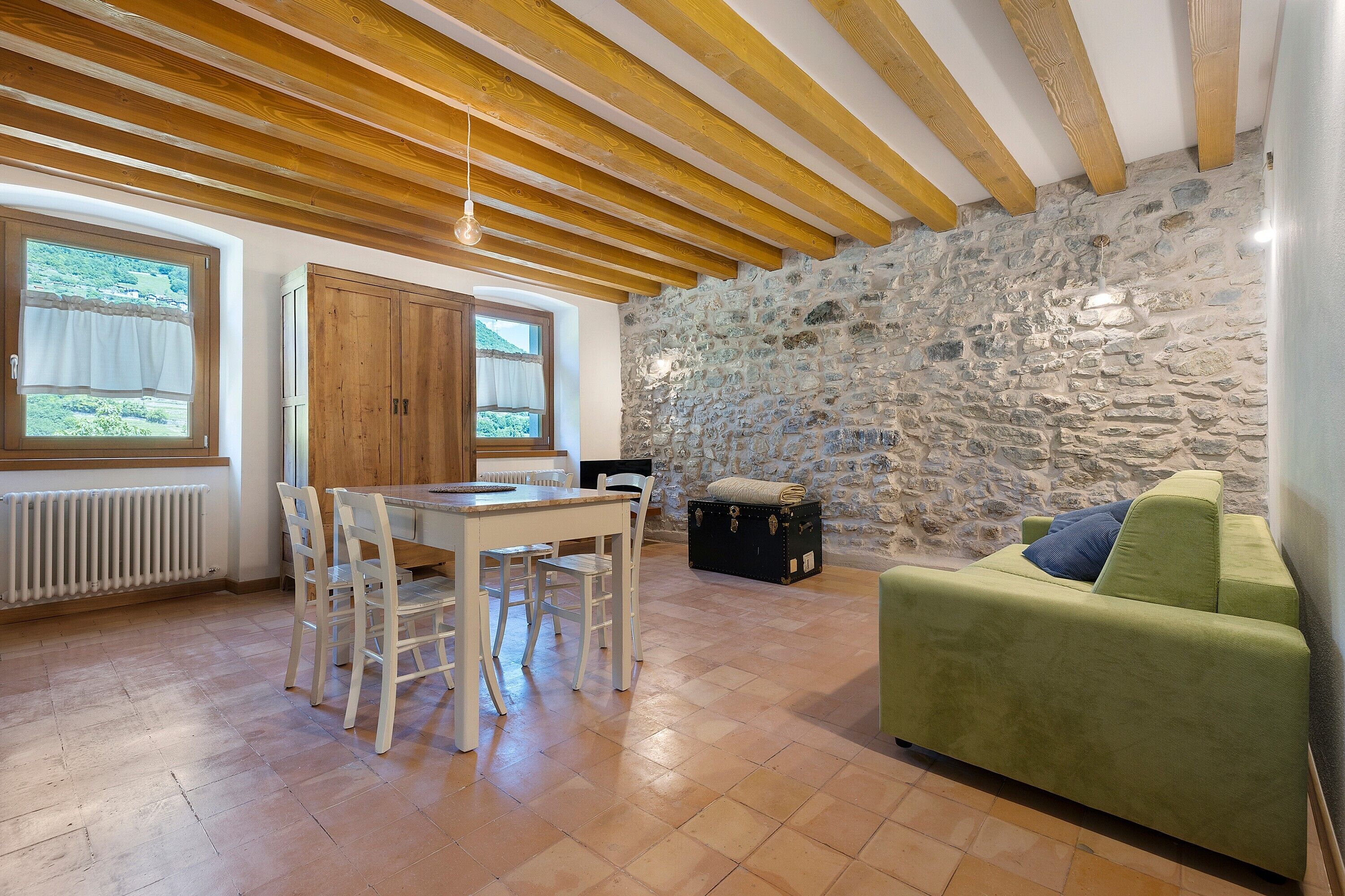 Une pièce avec un mur en pierre, des poutres en bois, un canapé vert, une table avec des chaises et une fenêtre donnant sur un paysage agréable.