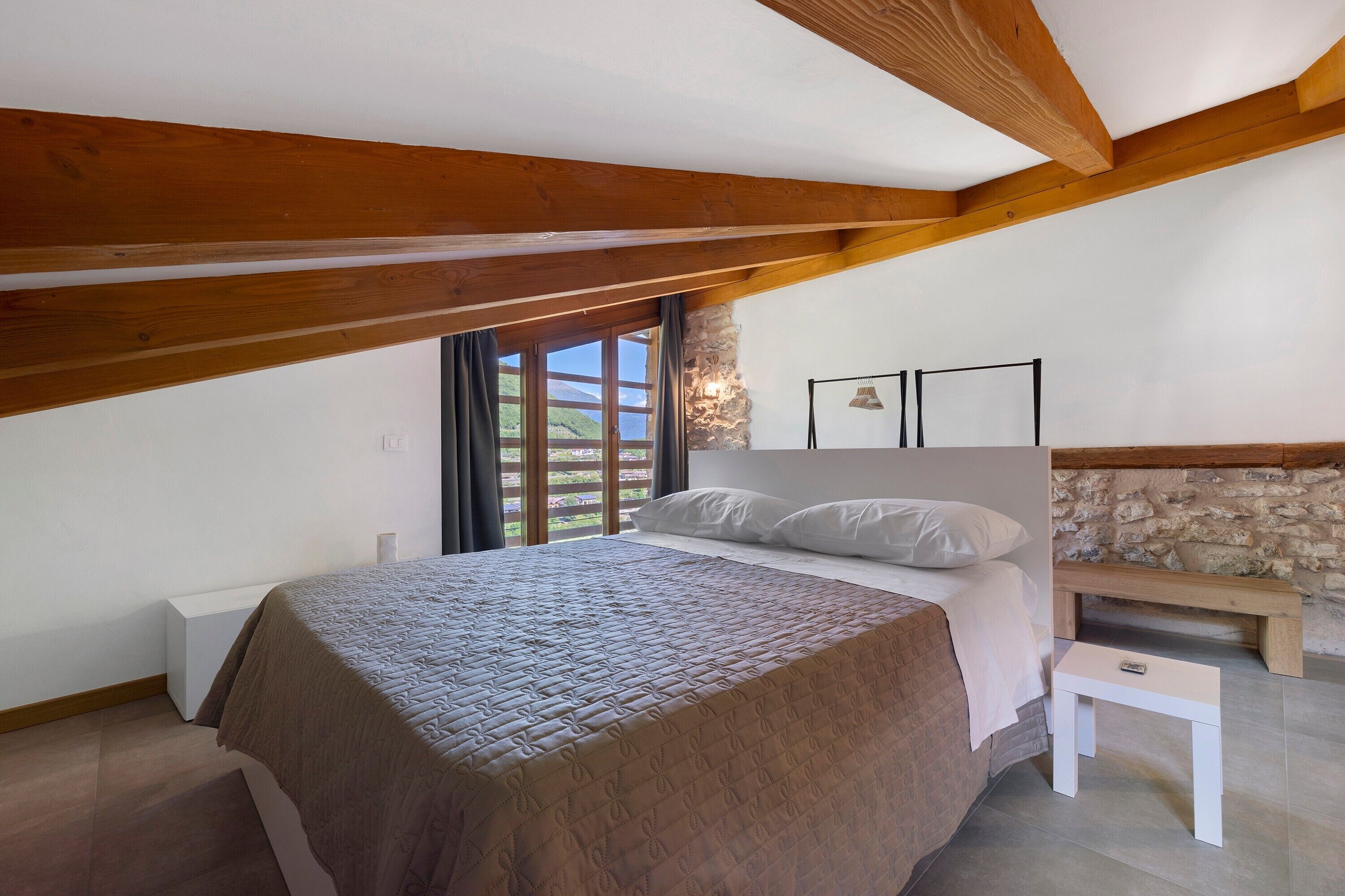 Une chambre à coucher avec un lit, une table de chevet, un banc, un mur en pierre et une fenêtre avec des rideaux.