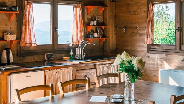L’intérieur d’un chalet en bois, avec une table à manger, des chaises, un coin cuisine et une vue par les fenêtres.
