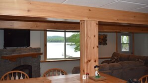 A wooden dining table with chairs, a stone fireplace, a television on a stone shelf, a large window with a view of a lake, and a brown sofa with patterned cushions. - Waterfront lake house with private beach and dock with hot  tub on Paradox Lake. (Schroon Lake)