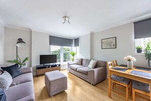 Ein modernes Wohnzimmer mit einer Couch, Hocker, Esstisch und Fernseher.