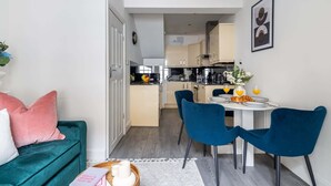 Un espace de vie compact avec un canapé turquoise, un coin repas avec des chaises bleues, une cuisine en arrière-plan et un tableau encadré au mur.