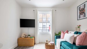 Un salon avec un canapé de couleur turquoise, un téléviseur à écran plat, une table d’appoint en bois et une fenêtre avec des rideaux.