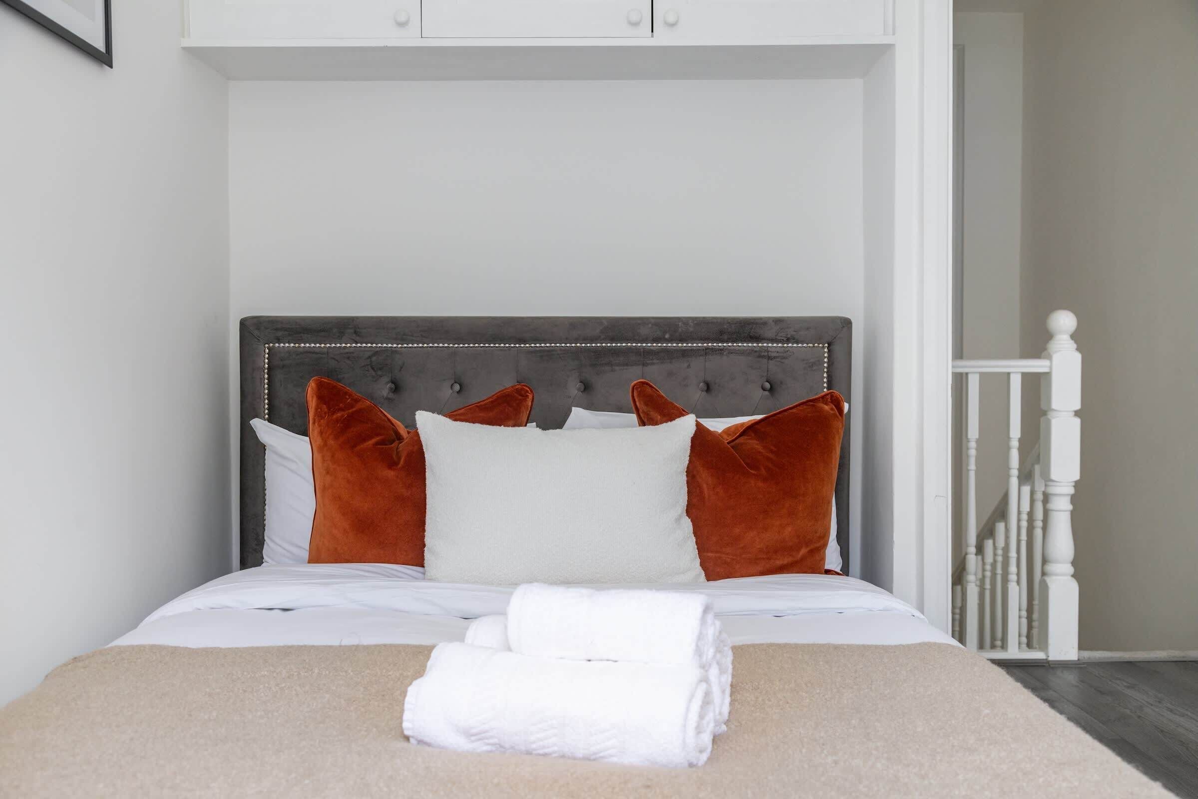 A neatly made bed with a grey upholstered headboard, white and orange pillows, and a folded white towel on the bed.