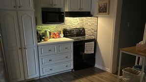 A compact kitchen with white cabinets, a black stove, and a microwave. There is a dining table with chairs and a trash can. - Entire Apartment in Bedford (Bedford)