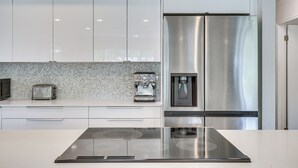 A modern kitchen with white cabinets, a stainless steel refrigerator, a coffee machine, and a built-in stovetop. - Beautiful, Calm, Modern Home with a Large Playroom (Hallandale Beach)