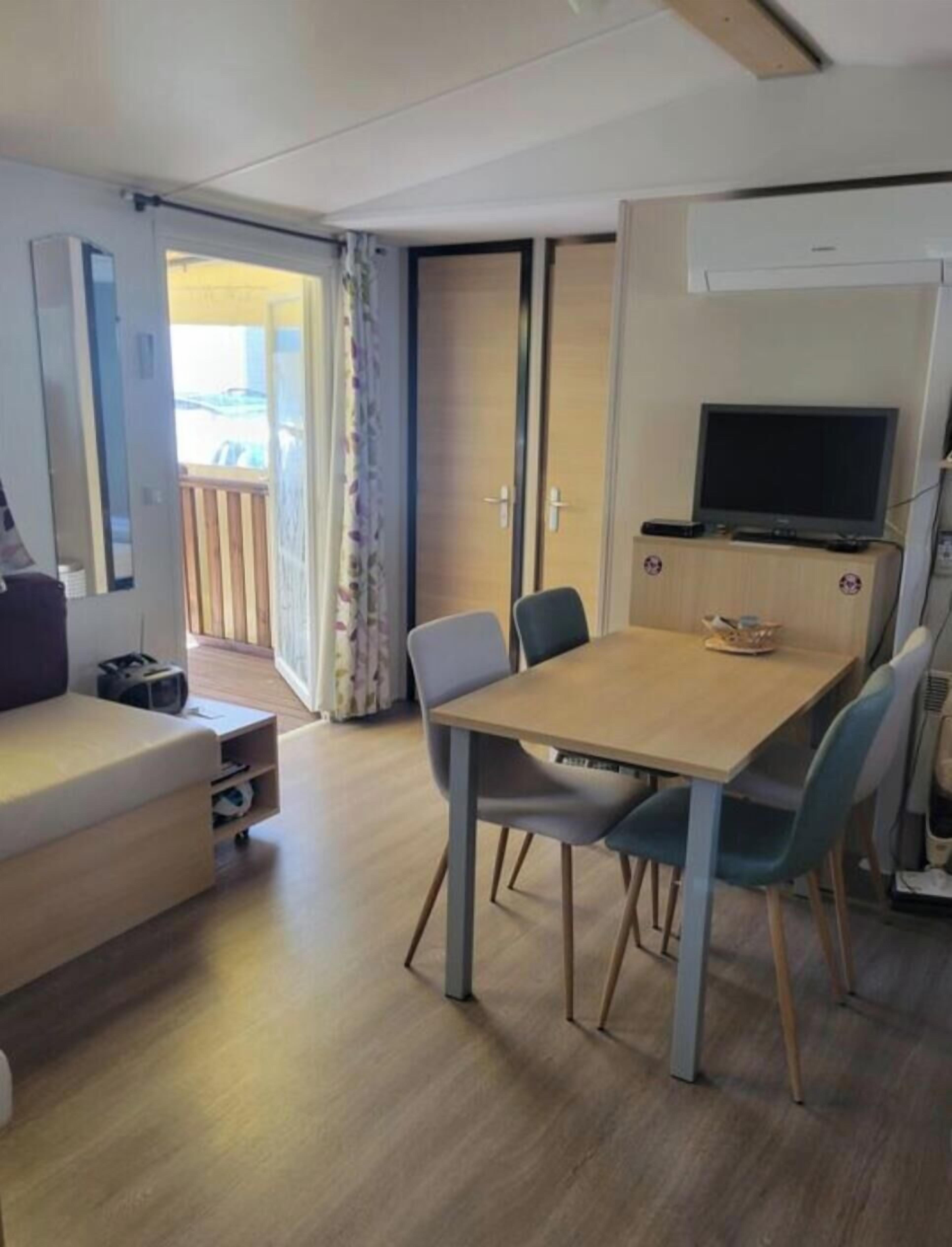 A room with a wooden dining table, chairs, a sofa, a TV, and an air conditioning unit.