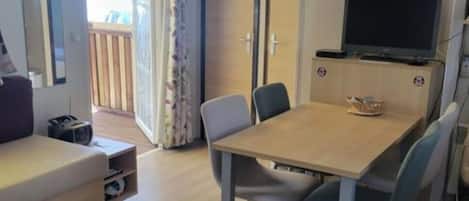A room with a wooden dining table, chairs, a sofa, a TV, and an air conditioning unit.