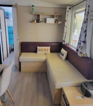 A compact living space with a sofa, a small table, a shelf with toiletries, and a window with curtains. - Mobile home in a 4-star campsite (Aureilhan)