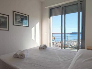 Une chambre d’hôtel avec un lit, deux serviettes enroulées, une fenêtre donnant sur la mer et les bâtiments, et deux tableaux encadrés accrochés au mur.