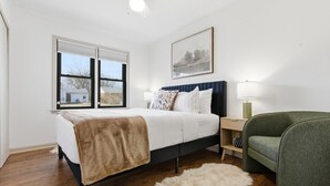 Una camera da letto con un letto, un comodino, una sedia, una finestra e un quadro appeso al muro.
