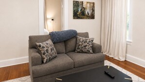 Een woonkamer met een bank, twee gestreepte kussens, een plaid, een zwarte salontafel, een afstandsbediening en een schilderij aan de muur.