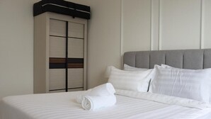 En pænt redt seng med hvide sengetøj og et sammenrullet håndklæde, en grå polstret sengegavl og et lukket skab med et sort-hvidt design.