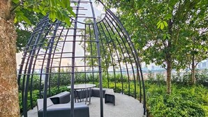 Et cirkulært udendørs siddeområde med en metalpergola, omgivet af grønt og træer.