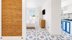 Une chambre d’hôtel moderne avec un lit, un panneau mural en bois, une télévision, un coin cuisine avec des placards bleus et des carreaux de sol à motifs.
