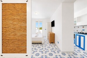 A modern hotel room with a bed, a wooden wall panel, a TV, a kitchen area with blue cabinets, and patterned floor tiles.