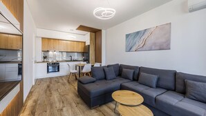 A modern living room with a sectional sofa, wooden flooring, a dining area, and a kitchen in the background. - Adriatic Escape Apartment | Steps from the Beach by PikHost (Lezha)