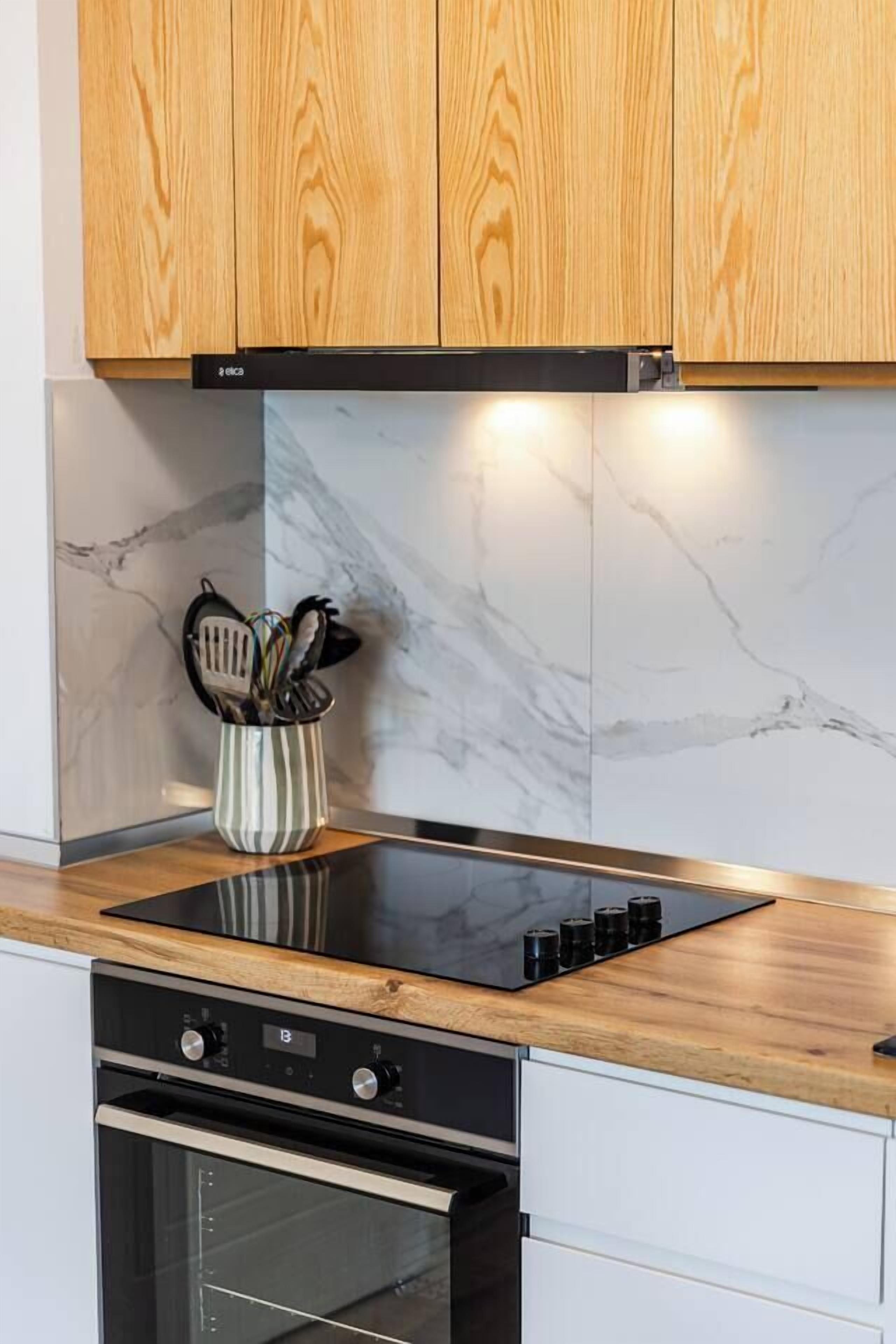 A kitchen with wooden cabinets, a black induction cooktop, a stainless steel oven, and a marble backsplash.