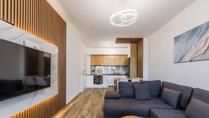 A modern living room with a flat-screen TV, a dark sofa, a wooden coffee table, and a kitchen in the background. - Adriatic Escape Apartment | Steps from the Beach by PikHost (Lezha)
