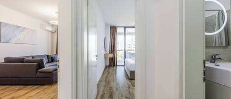 A modern apartment with a wooden floor, a living area with a sofa, a bedroom with a bed, and a bathroom with a sink and mirror.