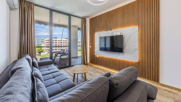 A modern living room with a sofa, a small round table, a TV mounted on a wooden panel wall, and a balcony with a view of buildings. - Adriatic Escape Apartment | Steps from the Beach by PikHost (Lezha)