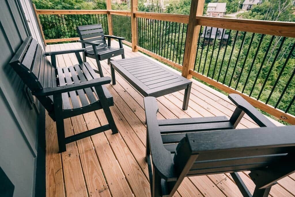A wooden deck with two black chairs and a small table, surrounded by a wooden railing and overlooking a green landscape.