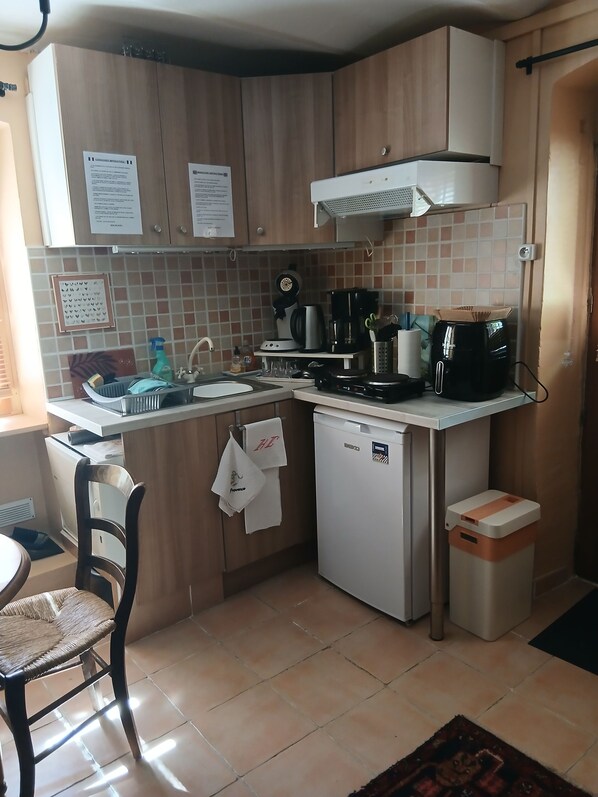 A compact kitchen with a sink, stove, and coffee maker. There are cabinets, a small refrigerator, and a trash bin.