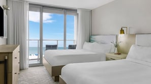 A hotel room with two beds, a desk, a chair, a nightstand, and a large window with a view of the ocean.